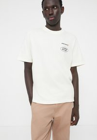Hvit bomull T-skjorte med korte ermer, rund hals, og svart brodert logo på brystet; kombinert med lyse beige bukser.