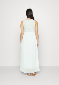 Vestido maxi verde menta com costas rendadas, cintura franzida e saia fluida, feito de um tecido leve e macio. Sandálias visíveis na parte inferior.