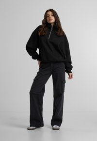 Svart fleece-pullover med halvnagskrage og lav skuldersøm, kombinert med svarte cargo-bukser som har sidelommer og vide bein.