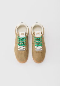 Et par tan semskede sneakers med grønne og hvite snorer, hvit innvendig foring, og "Steve Madden" metallmerker på snorene, lagt side om side.