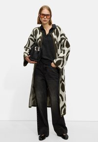 Long knit cardigan in muted green and cream leaf pattern, layered over a dark grey top, paired with black wide-leg jeans and a small black bag.