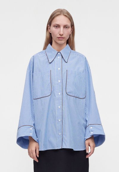Femme portant une chemise oversize à rayures bleues et blanches avec de grandes poches poitrine et des poignets larges, debout devant un fond uni.