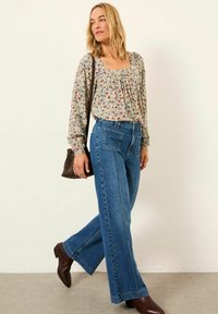 Woman wearing floral long-sleeve blouse, high-waisted wide-leg blue jeans, brown boots, and carrying a brown shoulder bag, walking indoors.