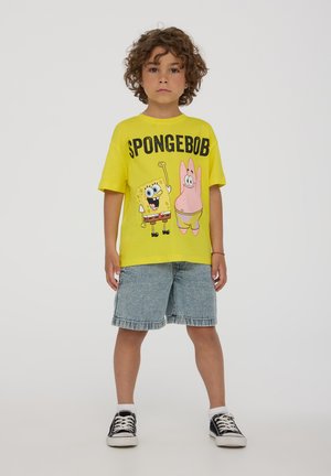 T-shirt di cotone gialla con personaggi dei cartoni animati SpongeBob e Patrick, abbinata a pantaloni shorts di jeans e sneakers nere.