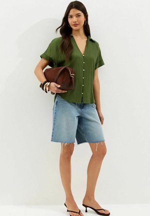 Kobieta w zielonej koszuli na krótki rękaw z guzikami i przetartych szortach dżinsowych do kolan, trzyma brązową torbę, czarne szpilki i nosi bransoletki.