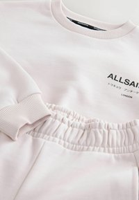 Jasnoróżowy dres z sweatshirtu i spodni z prążkowanymi mankietami, elastycznym pasem i napisem "ALLSAINTS LONDON" w japońskich znakach na piersi.