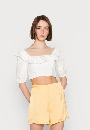 Blusa branca cropped com acabamento em renda de olho e mangas bufantes, combinada com calções amarelos de cetim com textura suave e um corte bem ajustado.