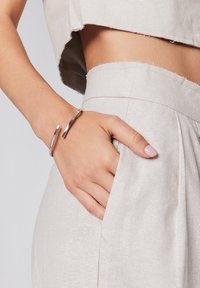 Srebrna metalowa bransoleta na nadgarstek, gładkie wykończenie, otwarty design z delikatnymi krzywiznami, noszona na dłoni spoczywającej w kieszeni jasnobeżowych spodni.