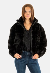 Veste bomber en fausse fourrure noire avec un col montant, fermeture éclair à l'avant et texture matelassée, associée à une chemise noire à pois et un jean bleu.