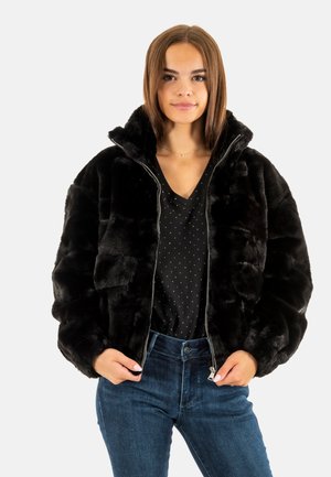 Giacca bomber in ecopelliccia nera con colletto alto, zip frontale e texture trapuntata, abbinata a una camicia nera a pois e jeans blu.