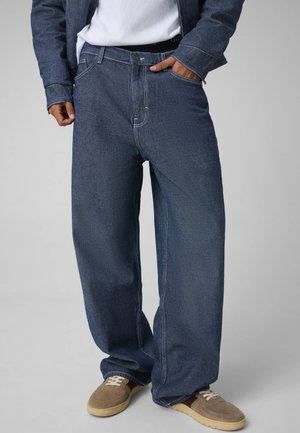 Modèle portant un jean ample bleu foncé, une veste en jean, une chemise blanche et des baskets marron ; une main repose dans une poche du jean.