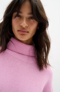 Maglione a collo alto rosa con collare a coste, texture morbida e vestibilità loose. Il viso del modello è parzialmente visibile con capelli scuri e mossi.