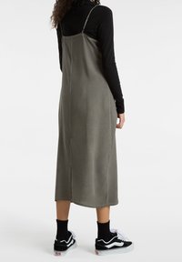 Vestido midi de tela verde oliva con tirantes ajustables, corte recto y una abertura lateral. Combinado con zapatillas negras y calcetines hasta el tobillo.