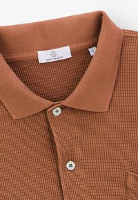 Camisa polo de textura waffle marrón con collar acanalado, placket de dos botones y detalles cosidos; incluye una etiqueta de marca debajo del collar.