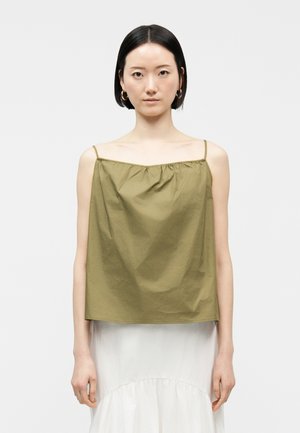 Γυναίκα με κοντά μαύρα μαλλιά που φοράει χρυσούς κρίκους στο αυτί, ένα αμάνικο τοπ πράσινο ελιάς με λεπτές τιράντες και μια λευκή φούστα.