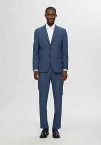 Costume bleu sur mesure en tissu texturé, avec un revers cranté, deux boutons et un pantalon assorti. Porté avec une chemise blanche et des chaussures noires.