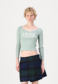 Šviesiai žalia ilgomis rankovėmis trumpa palaidinė su nėriniais ir balta užrašu "1998 SAN DIEGO". Papildyta mėlynai žaliais langeliais mini sijonu.