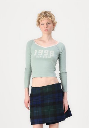 Lys grønn langermet crop topp med blonder og hvit "1998 SAN DIEGO" tekst. Kombinert med en miniskjørt i blå og grønn rute.