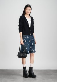 Cardigan nero lavorato a maglia con dettagli sui bottoni, pantaloni corti in denim ricamati con fiori bianchi, borsa a tracolla nera trapuntata e stivali con lacci e plateau.