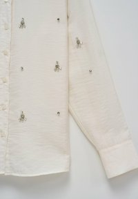 Chemise blanche à manches longues avec boutonnière sur le devant et petites décorations en cristal dispersées en forme florale et ronde.