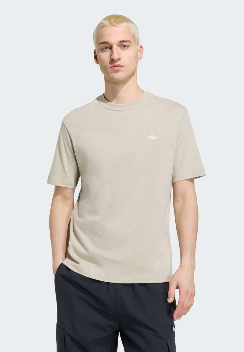 Jonge man met kort blond haar, die een beige Adidas T-shirt en zwarte cargobroek draagt, staat met één hand in zijn zak.