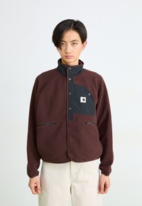 Persona joven con cabello corto y negro, vistiendo una chaqueta de forro polar marrón con un bolsillo negro en el pecho y pantalones beige, de pie frente a un fondo blanco liso.