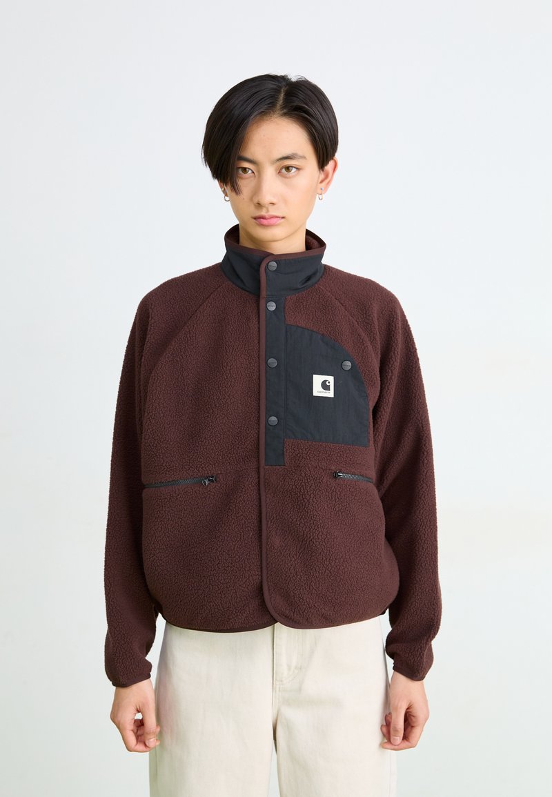 Persona joven con cabello corto y negro, vistiendo una chaqueta de forro polar marrón con un bolsillo negro en el pecho y pantalones beige, de pie frente a un fondo blanco liso.
