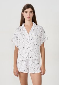 Jonge vrouw in een witte korte mouwen knoopjespajama en bijpassende korte broek met kleine roze en zwarte hartjespatronen, staande tegen een effen achtergrond.