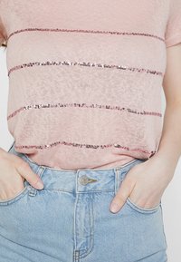 Jasnoróżowy sweter z krótkim rękawem ozdobiony poziomymi paskami z cekinów. Połączony z wysokim stanem jasnoniebieskich jeansowych szortów.