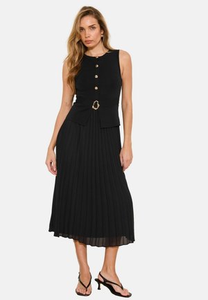 Žena má na sebe čierne plisované midi šaty bez rukávov so zlatými gombíkmi a detailom v tvare krúžku v páse, skombinované s čiernymi sandálmi na podpätku.
