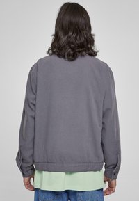 Šedá bavlněná bunda s volným střihem, s žebrovaným lemem, dlouhými rukávy a vrstveným zeleným tričkem pod ní.