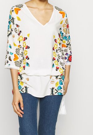 Femme portant un chemisier blanc à col en V avec des motifs colorés de papillons et floraux et des manches trois-quarts, associé à un jean bleu.