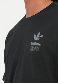 Juodas medvilnės T-marškinėlis su pilku Adidas logotipu ir grafikų dizainu krūtinėje. Yra klasikinis apvalus kaklas ir trumpomis rankovėmis.