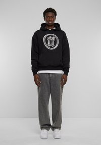 Felpa nera con cappuccio e grande logo argentato, abbinata a jeans grigi e sneakers bianche. Presenta un look stratificato e casual.