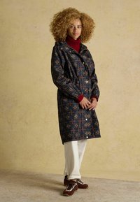 Garais, rakstainais tumši zilais mētelis ar elastīgu jostasvietu, kapuci un sarkaniem piedurknēm; valkāts virs sarkanas turtleneck blūzes un apvienots ar baltām biksēm un brūnām kurpēm.