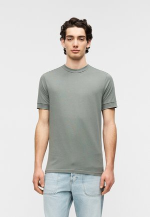 Fitted short-sleeve t-shirt in muted green cotton, featuring a ribbed crew neckline and rolled sleeves. Worn with light-wash jeans.
