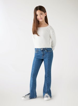 Ragazza giovane con lunghi capelli castani che indossa un maglione bianco con decorazioni, jeans blu con spaccature frontali e sneakers argentate, in piedi su sfondo bianco.