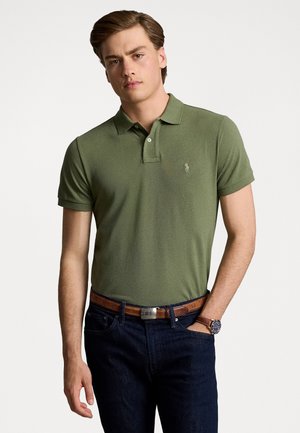 Camisa de polo verde con cuello, mangas cortas y un logo en el pecho. Combinada con unos jeans de mezclilla oscuros y un cinturón marrón.