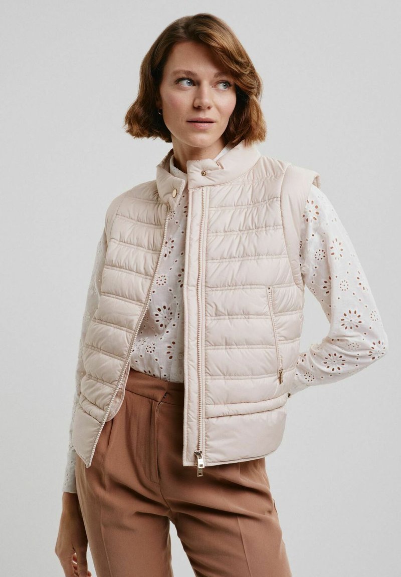 Beżowa pikowana kamizelka puchowa bez rękawów z wysokim kołnierzem, zamkiem błyskawicznym z przodu i dwoma bocznymi kieszeniami. Nosi ją nad białą bluzką z wyhaftowanymi otworami.