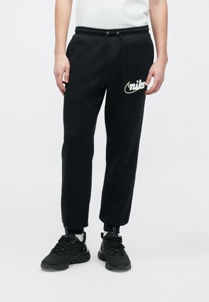 Personne portant un pantalon de survêtement Nike noir avec un logo blanc sur la cuisse gauche, des baskets noires et un t-shirt blanc, debout sur un fond blanc uni.