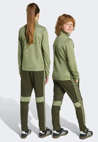 Olivově zelená sportovní bunda s vysokým límcem v kombinaci s odpovídajícími zúženými kalhotami s bočními pruhy a akcenty na kolenou.