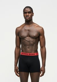 Homme musclé debout, portant un boxer noir avec une ceinture rouge marquée "BJÖRN BORG", fond gris neutre.