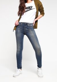Jeans ajustados en denim azul oscuro, con un corte slim, ligero desgaste, camiseta blanca con el logo de Nike y chaqueta con cremallera verde olivo, zapatillas blancas.