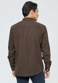 Chemise à carreaux brune en coton, dotée d'un col classique, de manches longues et de poignets boutonnés. La vue de dos montre la longueur et la coupe de la chemise.