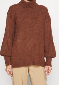 Brązowy, dziergany sweter z golfem o luźnym kroju i bufiastych rękawach. Szczegółowe prążkowanie przy dekolcie i mankietach. W zestawieniu z beżowymi spodniami.