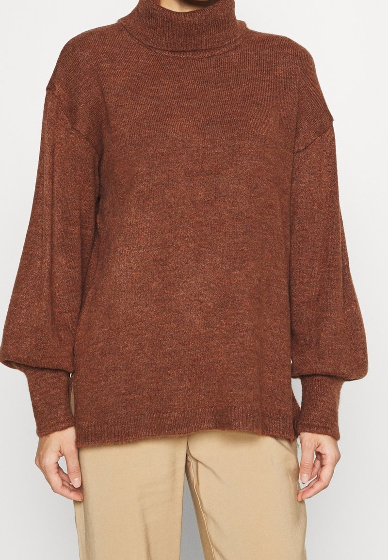 Brązowy, dziergany sweter z golfem o luźnym kroju i bufiastych rękawach. Szczegółowe prążkowanie przy dekolcie i mankietach. W zestawieniu z beżowymi spodniami.