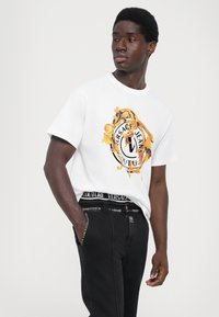 Hvit bomull T-skjorte med et gull- og svart brodert Versace-logo. Kombinert med mørk denimjeans, akkompagnert av et merkevarebelte.