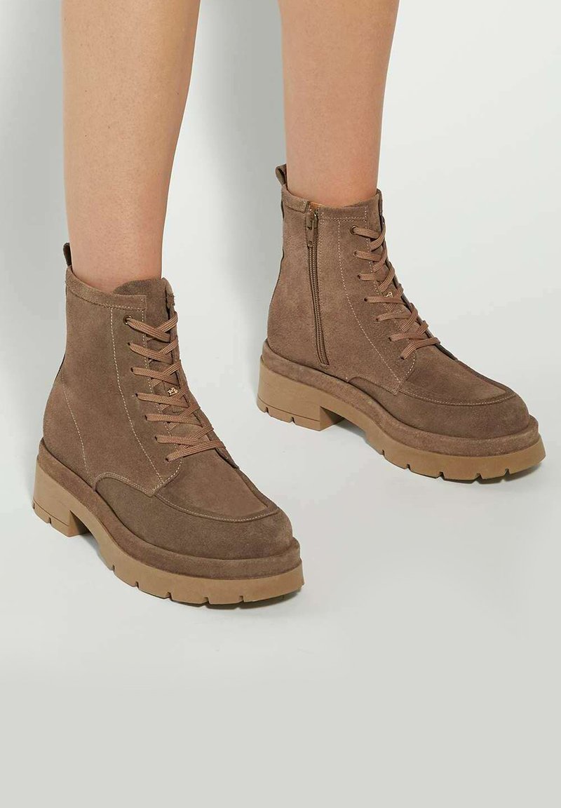Brown suede ankle boots with laces and side zipper. Chunky beige sole and subtle stitching details enhance the design. Flat heel.