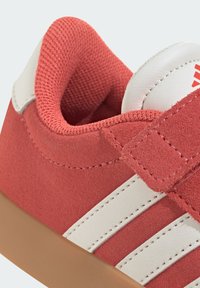 Červené semišové topánky s bielymi koženými pruhmi, polstrovaným golierom a béžovou gumovou podrážkou s viditeľnými švami.