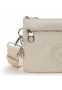 Beige korsväska med silverfärgad dragkedja, avtagbar bred axelrem och liten silverfärgad apa nyckelring charm på vit bakgrund.